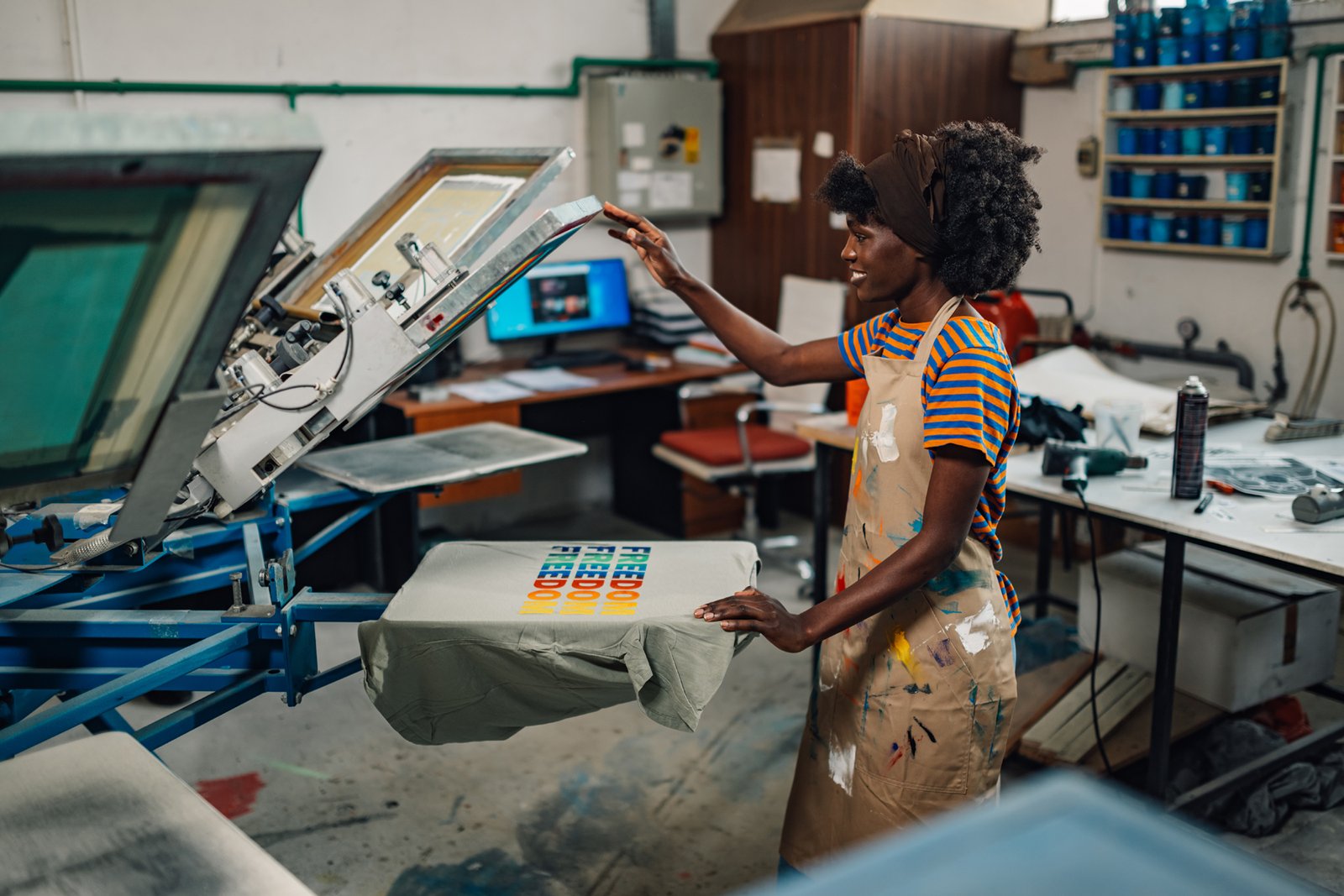 Side view of smiling african american graphic technology expert standing at printing workshop and finishing printing on a silk screen manual printing machine.Multicultural employee working at facility