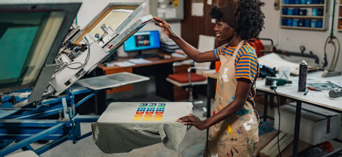 Side view of smiling african american graphic technology expert standing at printing workshop and finishing printing on a silk screen manual printing machine.Multicultural employee working at facility